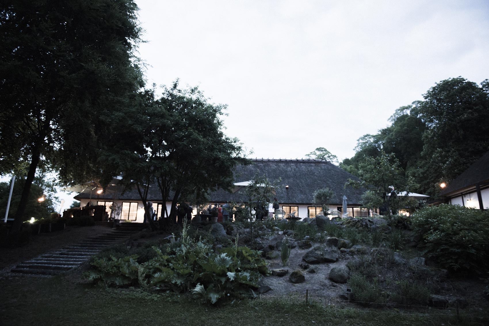 Gæsterne på terrassen ved Restaurant Herthadalen i aftenlys, omgivet af smuk grøn natur under Kathrine og Michaels bryllupsfest, fotograferet af To the Moon and Back.